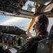 A military pilot is seated in the cockpit of a large aircraft, likely a tanker or transport plane, during flight. The pilot is wearing a dark flight suit, a Bose aviation headset with boom microphone, and has a squadron patch visible on his shoulder. The cockpit features an extensive array of analog and digital instrumentation panels.