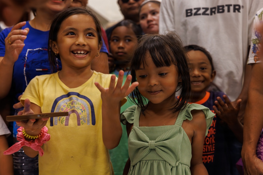 A group of children and adults gathered together, with two young girls prominently featured in the foreground. The girl on the left wears a yellow t-shirt with a rainbow design and is smiling broadly while holding a smartphone. The girl on the right wears a green dress with a bow and is waving at the camera.