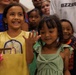 A group of children and adults gathered together, with two young girls prominently featured in the foreground. The girl on the left wears a yellow t-shirt with a rainbow design and is smiling broadly while holding a smartphone. The girl on the right wears a green dress with a bow and is waving at the camera.