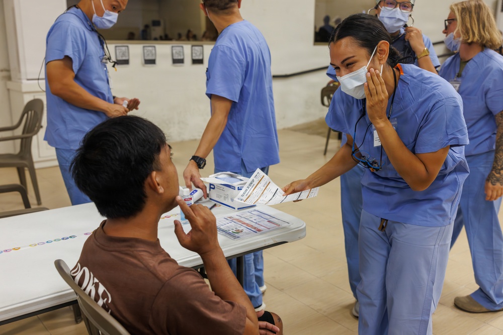 A medical or health outreach event where healthcare workers in blue scrubs and surgical masks are attending to patients or community members. A female healthcare worker is handing informational pamphlets/brochures to a seated young male patient while another item (possibly a thermometer or medical device) is being used near his face. Multiple healthcare personnel are visible in the background attending to other individuals.