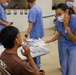 A medical or health outreach event where healthcare workers in blue scrubs and surgical masks are attending to patients or community members. A female healthcare worker is handing informational pamphlets/brochures to a seated young male patient while another item (possibly a thermometer or medical device) is being used near his face. Multiple healthcare personnel are visible in the background attending to other individuals.