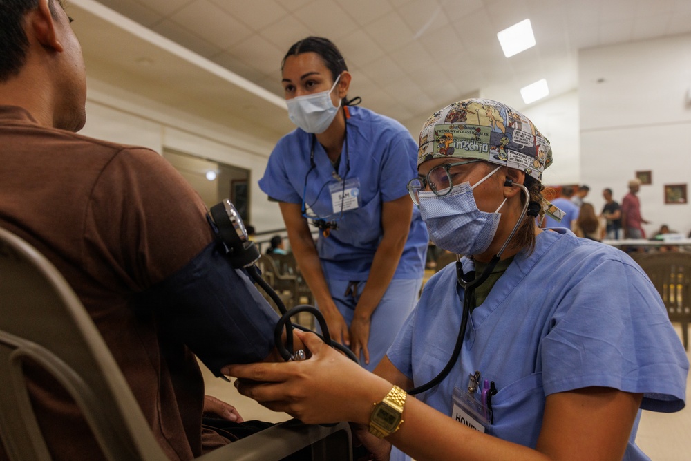Two healthcare workers in blue scrubs and surgical masks are attending to a patient in what appears to be a community health clinic or medical outreach event. The foreground worker is taking the patient's blood pressure using a sphygmomanometer and stethoscope, while a second provider observes. The setting appears to be a large indoor hall with multiple people visible in the background.