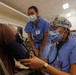 Two healthcare workers in blue scrubs and surgical masks are attending to a patient in what appears to be a community health clinic or medical outreach event. The foreground worker is taking the patient's blood pressure using a sphygmomanometer and stethoscope, while a second provider observes. The setting appears to be a large indoor hall with multiple people visible in the background.