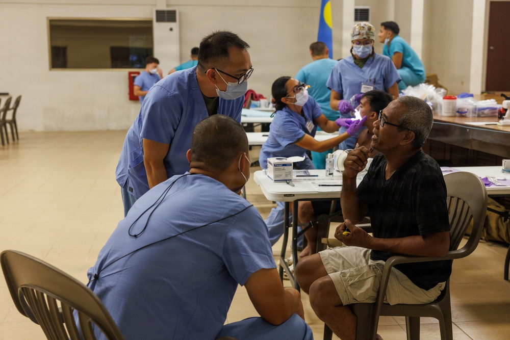 A medical outreach or field clinic setting where multiple healthcare workers in blue scrubs are attending to patients in what appears to be a community hall or gymnasium. At least two patients are being examined simultaneously, with dental or oral examinations visible in the background. The setting suggests a mobile medical mission or humanitarian health event.
