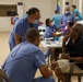 A medical outreach or field clinic setting where multiple healthcare workers in blue scrubs are attending to patients in what appears to be a community hall or gymnasium. At least two patients are being examined simultaneously, with dental or oral examinations visible in the background. The setting suggests a mobile medical mission or humanitarian health event.