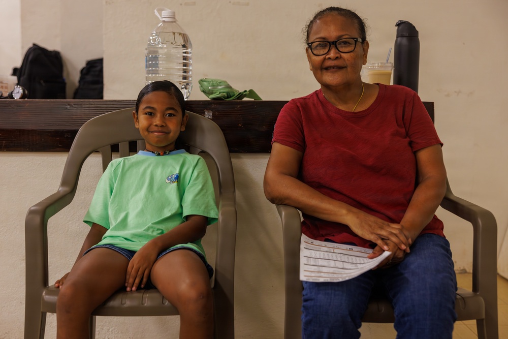 Two individuals, a young child and an older woman, are seated side by side on gray plastic chairs against a light-colored wall. The child is wearing a green t-shirt and denim shorts, while the older woman wears a red t-shirt, jeans, and glasses. Both are facing the camera and appear relaxed in what looks like a waiting area.