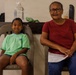 Two individuals, a young child and an older woman, are seated side by side on gray plastic chairs against a light-colored wall. The child is wearing a green t-shirt and denim shorts, while the older woman wears a red t-shirt, jeans, and glasses. Both are facing the camera and appear relaxed in what looks like a waiting area.