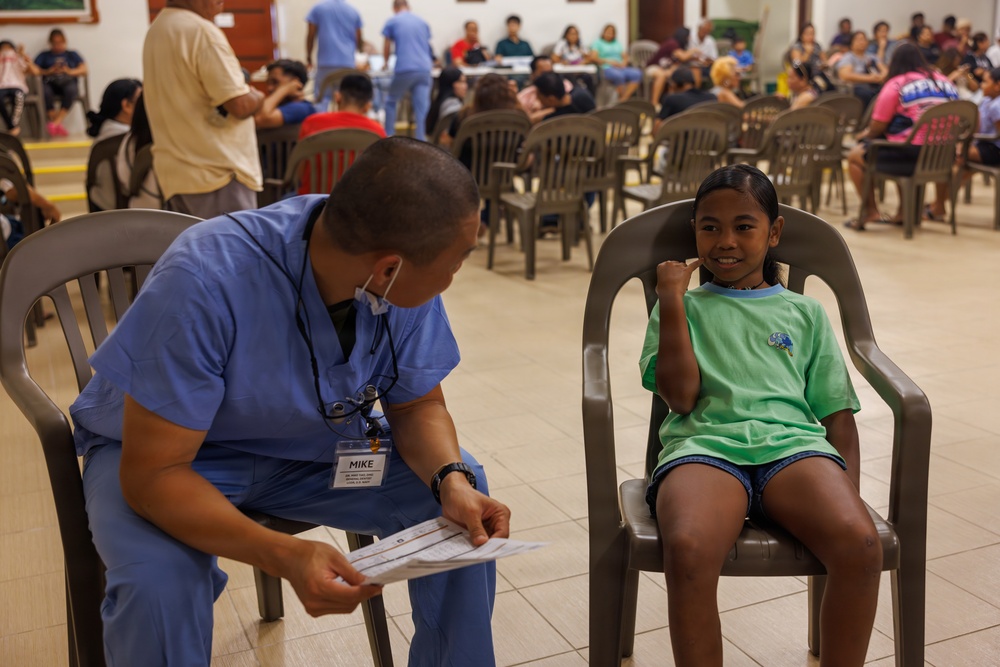 A healthcare worker named Mike, wearing blue scrubs and a name badge, is consulting with a young girl seated in a plastic chair in what appears to be a community health clinic or medical outreach event. The girl, wearing a green t-shirt, is smiling and making a fist gesture, suggesting a positive interaction. The background shows a large hall filled with many people waiting, indicating a high-volume medical outreach or vaccination event.