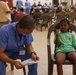 A healthcare worker named Mike, wearing blue scrubs and a name badge, is consulting with a young girl seated in a plastic chair in what appears to be a community health clinic or medical outreach event. The girl, wearing a green t-shirt, is smiling and making a fist gesture, suggesting a positive interaction. The background shows a large hall filled with many people waiting, indicating a high-volume medical outreach or vaccination event.