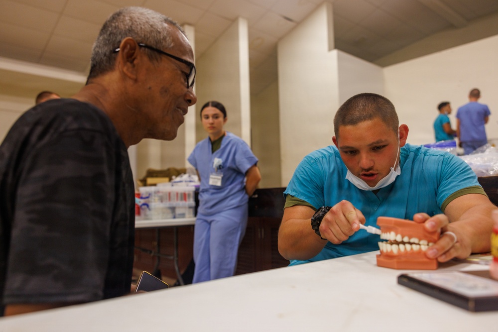 A dental health education session is taking place in what appears to be a community health clinic or outreach event. A healthcare worker in teal scrubs is demonstrating proper tooth brushing technique using a dental model to an older male patient. Additional medical staff in scrubs are visible in the background, along with supplies on tables.
