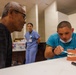 A dental health education session is taking place in what appears to be a community health clinic or outreach event. A healthcare worker in teal scrubs is demonstrating proper tooth brushing technique using a dental model to an older male patient. Additional medical staff in scrubs are visible in the background, along with supplies on tables.
