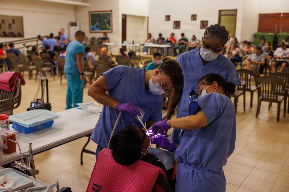 A dental outreach or medical mission event taking place in a large community hall or multipurpose room. Three dental professionals in blue scrubs and masks are performing a dental procedure on a seated patient wearing a pink bib. The background shows a waiting area filled with numerous patients, suggesting a high-volume free or low-cost dental clinic.