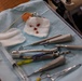 A dental procedure tray is shown post-operation, containing various used dental instruments laid out on a sterile blue drape. Gauze pads are visible, one of which contains small bloody tissue fragments, indicating a recent tooth extraction or oral surgery procedure. The tray includes a variety of stainless steel dental tools alongside a syringe and color-coded instruments.