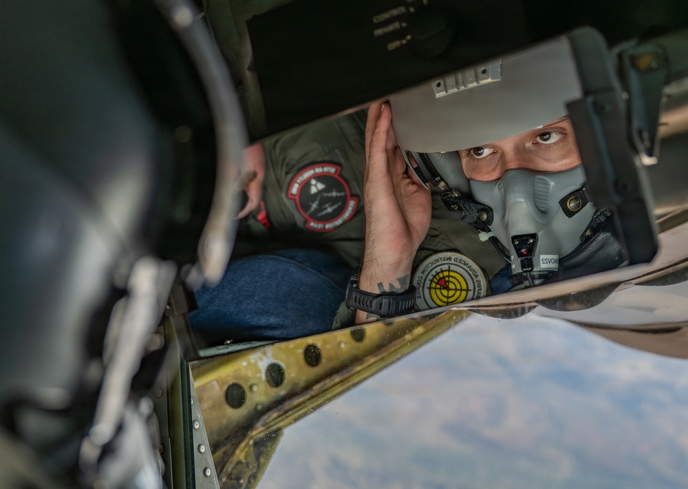 A military aviator wearing an oxygen mask and flight suit is visible through a narrow opening in an aircraft structure, appearing to inspect or check equipment while airborne. The person is wearing a green flight suit with unit patches, including one that reads 'Advanced Intelligence Course' with a radar/target symbol. The terrain visible below suggests the aircraft is at significant altitude.