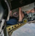 A military aviator wearing an oxygen mask and flight suit is visible through a narrow opening in an aircraft structure, appearing to inspect or check equipment while airborne. The person is wearing a green flight suit with unit patches, including one that reads 'Advanced Intelligence Course' with a radar/target symbol. The terrain visible below suggests the aircraft is at significant altitude.