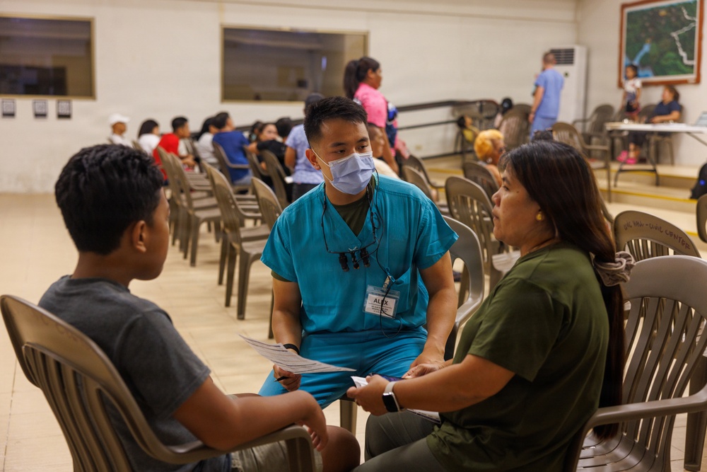 A healthcare worker wearing teal scrubs and a surgical mask, identified by a name tag reading 'ALEX', is seated and consulting with a woman in an olive green shirt and a young boy in a gray shirt. The setting appears to be a community health clinic or medical outreach event held in a large hall. Multiple people are visible in the background, seated in rows of plastic chairs, suggesting a waiting area.