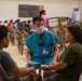 A healthcare worker wearing teal scrubs and a surgical mask, identified by a name tag reading 'ALEX', is seated and consulting with a woman in an olive green shirt and a young boy in a gray shirt. The setting appears to be a community health clinic or medical outreach event held in a large hall. Multiple people are visible in the background, seated in rows of plastic chairs, suggesting a waiting area.