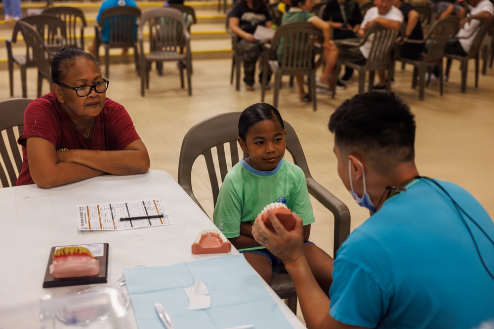 A dental health education session is taking place at what appears to be a community health outreach event. A dental professional in a blue scrub top and face mask is showing a dental model to a young child in a green shirt, while an older woman (likely a guardian) observes attentively. The setting is a large open hall with multiple people seated in the background, suggesting a organized community health fair.