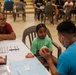 A dental health education session is taking place at what appears to be a community health outreach event. A dental professional in a blue scrub top and face mask is showing a dental model to a young child in a green shirt, while an older woman (likely a guardian) observes attentively. The setting is a large open hall with multiple people seated in the background, suggesting a organized community health fair.
