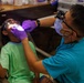 A dental professional is examining a young child's mouth in what appears to be a field or community dental clinic setting. The dentist is wearing protective gear including loupes, a face mask, and purple nitrile gloves, and is using a light to illuminate the child's oral cavity. The child, wearing a green t-shirt, is reclined in a chair with protective eyewear on.