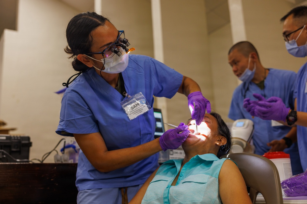 A dental procedure is being performed in a clinical setting. A dental professional identified by a name badge reading 'SAM' is treating a seated patient while using magnification loupes and a headlight. Two additional medical personnel in blue scrubs and purple gloves are present in the background.