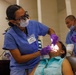 A dental procedure is being performed in a clinical setting. A dental professional identified by a name badge reading 'SAM' is treating a seated patient while using magnification loupes and a headlight. Two additional medical personnel in blue scrubs and purple gloves are present in the background.