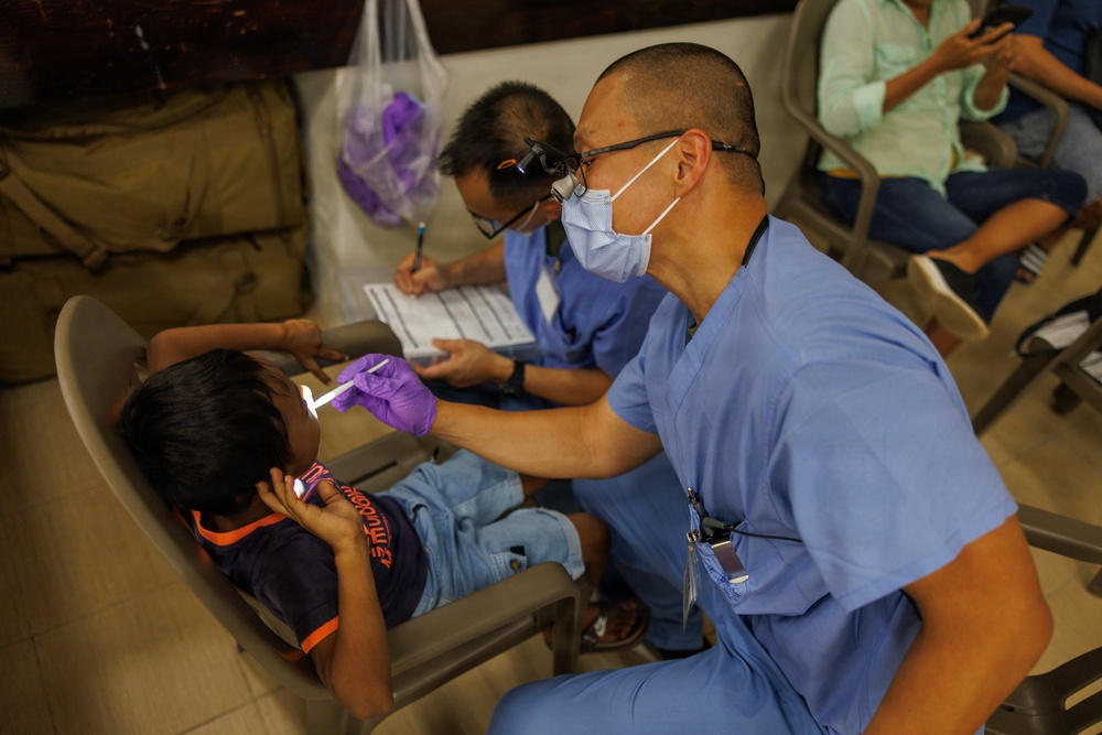 A dental professional in blue scrubs, surgical mask, and purple gloves examines a young child's mouth using a light instrument in what appears to be a field or community dental clinic. A second medical professional in the background is taking notes on a clipboard. Several people are visible waiting in chairs in the background.