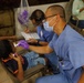 A dental professional in blue scrubs, surgical mask, and purple gloves examines a young child's mouth using a light instrument in what appears to be a field or community dental clinic. A second medical professional in the background is taking notes on a clipboard. Several people are visible waiting in chairs in the background.