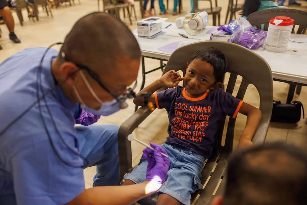 A young child is seated in a chair receiving a dental examination at what appears to be a community health clinic or outreach event. A dental professional wearing blue scrubs, a face mask, magnifying loupes, and purple gloves is performing the examination using a dental light. The child appears slightly apprehensive but is looking toward the camera with a cautious expression.