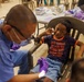 A young child is seated in a chair receiving a dental examination at what appears to be a community health clinic or outreach event. A dental professional wearing blue scrubs, a face mask, magnifying loupes, and purple gloves is performing the examination using a dental light. The child appears slightly apprehensive but is looking toward the camera with a cautious expression.