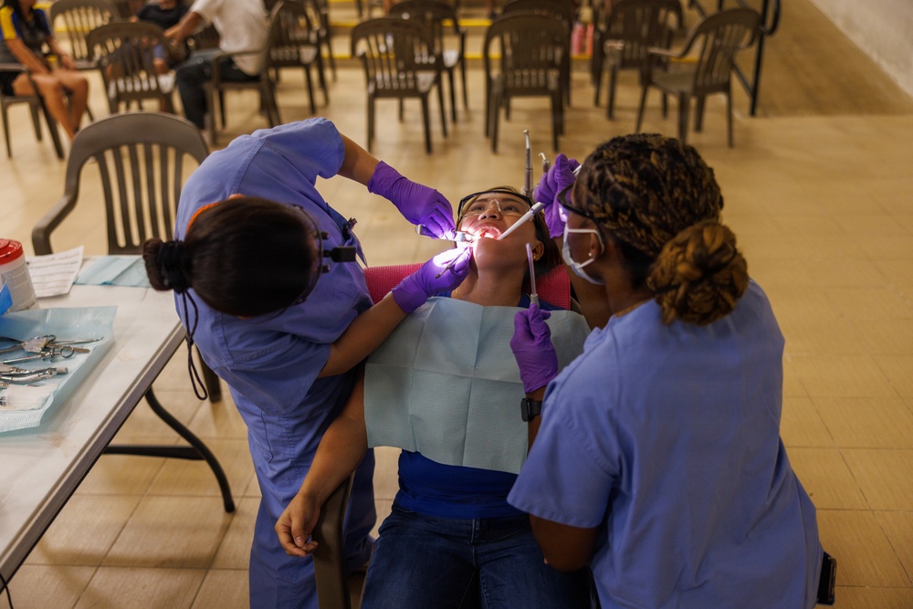 Two dental professionals in blue scrubs and purple gloves are performing a dental procedure on a patient reclined in a portable dental chair. The setting appears to be a temporary or community dental clinic, likely a medical outreach event, set up in a large open hall. The patient has a dental bib and protective eyewear while receiving treatment with illuminated dental instruments.