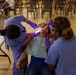 Two dental professionals in blue scrubs and purple gloves are performing a dental procedure on a patient reclined in a portable dental chair. The setting appears to be a temporary or community dental clinic, likely a medical outreach event, set up in a large open hall. The patient has a dental bib and protective eyewear while receiving treatment with illuminated dental instruments.