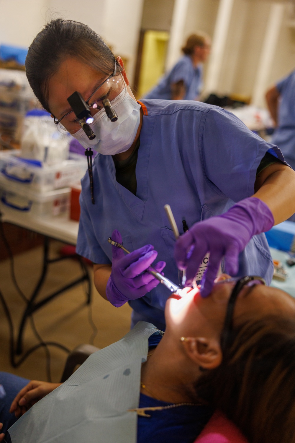A dental professional wearing blue scrubs, a surgical mask, magnifying loupes with an attached light, and purple nitrile gloves is performing a dental procedure on a reclined patient. The setting appears to be a community or volunteer dental clinic, evidenced by the informal environment with folding tables and multiple practitioners visible in the background. The patient is wearing a dental bib and has their mouth open while the dentist works with illuminated dental instruments.