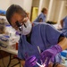 A dental professional wearing blue scrubs, a surgical mask, magnifying loupes with an attached light, and purple nitrile gloves is performing a dental procedure on a reclined patient. The setting appears to be a community or volunteer dental clinic, evidenced by the informal environment with folding tables and multiple practitioners visible in the background. The patient is wearing a dental bib and has their mouth open while the dentist works with illuminated dental instruments.
