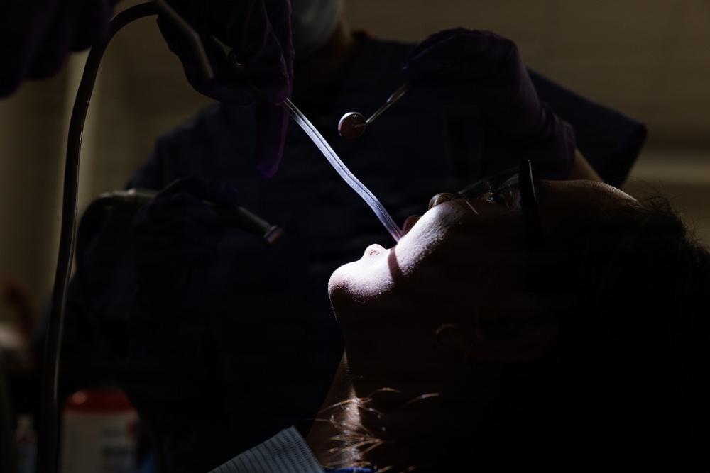 A dental procedure is being performed on a patient who is reclined in a dental chair. A dental professional wearing purple gloves is using instruments including what appears to be a dental mirror and suction tube. The image is dramatically lit with high contrast, leaving much of the scene in shadow.