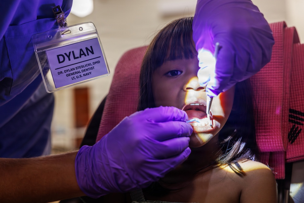 A U.S. Navy general dentist, Dr. Dylan Steslicki, DMD, is performing a dental examination on a young girl. The dentist is wearing purple nitrile gloves and using a dental light and instrument to examine the child's teeth. The child is seated in a dental chair with a red/pink drape visible in the background.