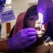 A U.S. Navy general dentist, Dr. Dylan Steslicki, DMD, is performing a dental examination on a young girl. The dentist is wearing purple nitrile gloves and using a dental light and instrument to examine the child's teeth. The child is seated in a dental chair with a red/pink drape visible in the background.