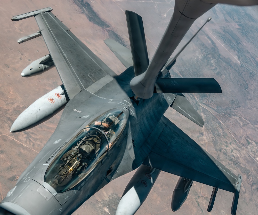 An aerial refueling operation is underway, with a fighter jet receiving fuel from a tanker aircraft via a boom refueling system. The fighter appears to be an F-16 Fighting Falcon, identifiable by its single-seat bubble canopy and delta wing configuration, bearing markings consistent with a Middle Eastern air force. The photograph is taken from the tanker's boom operator perspective, looking down at the receiver aircraft over an arid, desert landscape.
