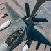 An aerial refueling operation is underway, with a fighter jet receiving fuel from a tanker aircraft via a boom refueling system. The fighter appears to be an F-16 Fighting Falcon, identifiable by its single-seat bubble canopy and delta wing configuration, bearing markings consistent with a Middle Eastern air force. The photograph is taken from the tanker's boom operator perspective, looking down at the receiver aircraft over an arid, desert landscape.