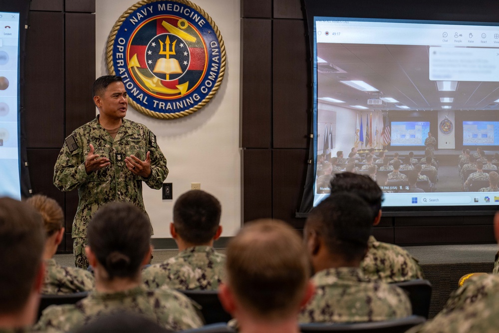 A U.S. Navy officer is delivering a presentation or briefing to a group of uniformed military personnel at the Navy Medicine Operational Training Command. The speaker stands in front of the command's official seal and a large display screen showing a video conference or live feed of another audience in a similar setting. The event appears to be a hybrid in-person and virtual training or briefing session.