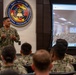 A U.S. Navy officer is delivering a presentation or briefing to a group of uniformed military personnel at the Navy Medicine Operational Training Command. The speaker stands in front of the command's official seal and a large display screen showing a video conference or live feed of another audience in a similar setting. The event appears to be a hybrid in-person and virtual training or briefing session.