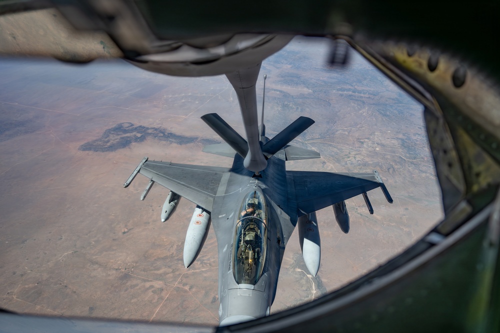An aerial refueling operation is captured from the boom operator's window of a tanker aircraft, showing a fighter jet receiving fuel mid-flight. The receiving aircraft appears to be an F-16 Fighting Falcon connected to the refueling boom of what is likely a KC-135 or similar tanker. The image is taken from a high altitude over an arid, semi-desert landscape.