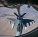 An aerial refueling operation is captured from the boom operator's window of a tanker aircraft, showing a fighter jet receiving fuel mid-flight. The receiving aircraft appears to be an F-16 Fighting Falcon connected to the refueling boom of what is likely a KC-135 or similar tanker. The image is taken from a high altitude over an arid, semi-desert landscape.