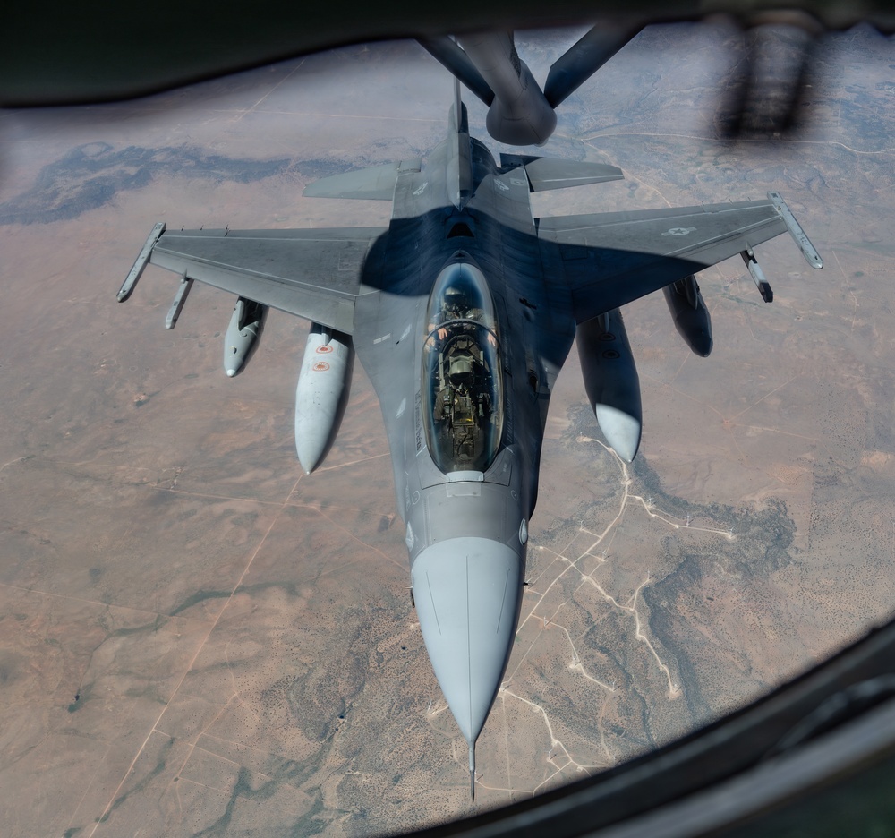 An aerial refueling operation is captured from the perspective of a tanker aircraft, showing a single-seat fighter jet receiving fuel via a boom refueling system. The fighter aircraft, identifiable as an F-16 Fighting Falcon, is flying in close proximity beneath the tanker over an arid, desert-like terrain. The photograph is taken from the tanker's boom operator window, providing a top-down forward view of the receiver aircraft.