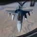 An aerial refueling operation is captured from the perspective of a tanker aircraft, showing a single-seat fighter jet receiving fuel via a boom refueling system. The fighter aircraft, identifiable as an F-16 Fighting Falcon, is flying in close proximity beneath the tanker over an arid, desert-like terrain. The photograph is taken from the tanker's boom operator window, providing a top-down forward view of the receiver aircraft.