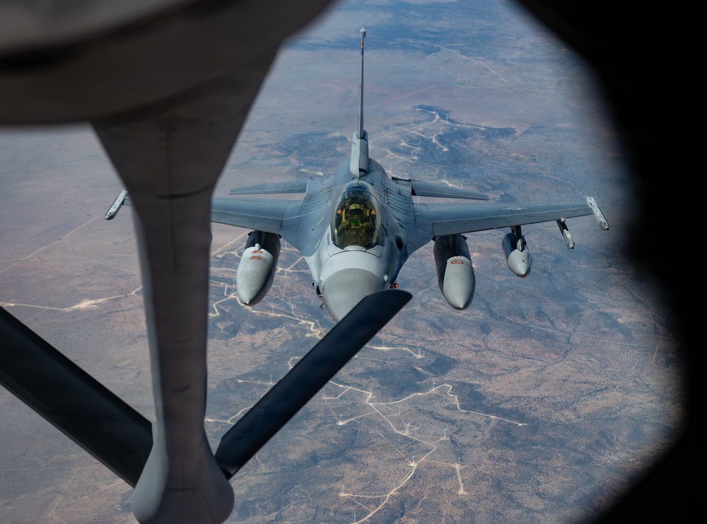 An aerial refueling operation is captured from the perspective of a tanker aircraft, showing an F-16 Fighting Falcon approaching the refueling boom. The fighter jet is photographed through the boom operator's window of the tanker, with an arid, sparsely populated landscape visible far below. The aircraft appears to be in a standard gray military paint scheme and is carrying external stores on its wing pylons.