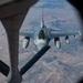 An aerial refueling operation is captured from the perspective of a tanker aircraft, showing an F-16 Fighting Falcon approaching the refueling boom. The fighter jet is photographed through the boom operator's window of the tanker, with an arid, sparsely populated landscape visible far below. The aircraft appears to be in a standard gray military paint scheme and is carrying external stores on its wing pylons.