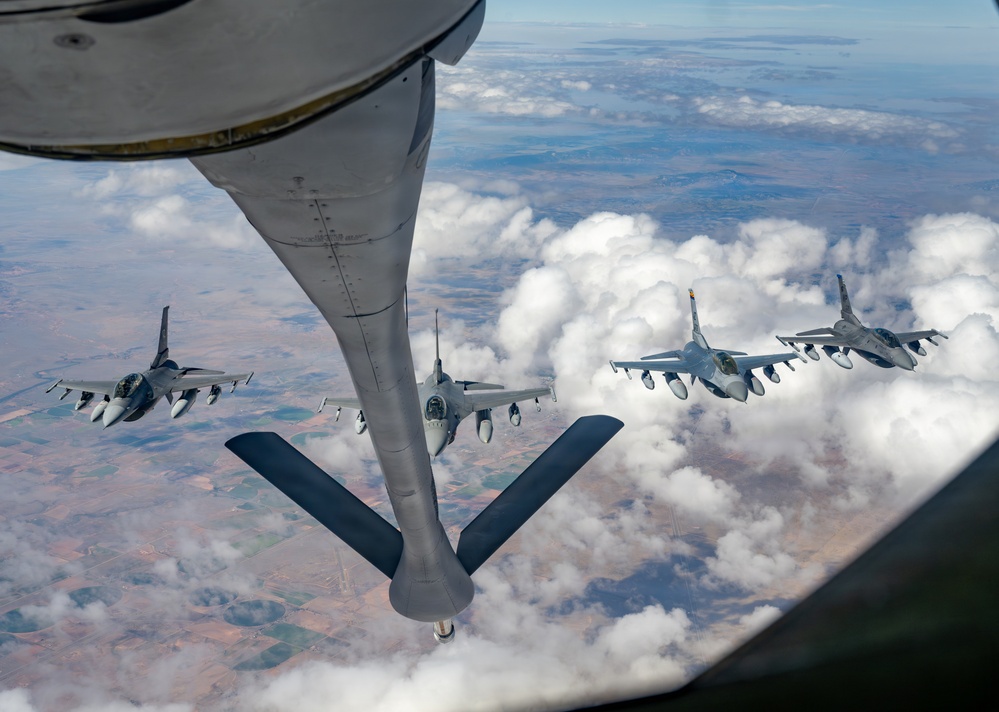 An aerial refueling operation is captured from the boom operator's station of a tanker aircraft, showing its refueling boom extended toward a receiver aircraft. Four F-16 Fighting Falcon jets are visible in formation, with one actively positioned for fuel transfer while others fly in escort positions. The image is taken at high altitude over a partially cloudy landscape with agricultural terrain visible below.