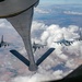 An aerial refueling operation is captured from the boom operator's station of a tanker aircraft, showing its refueling boom extended toward a receiver aircraft. Four F-16 Fighting Falcon jets are visible in formation, with one actively positioned for fuel transfer while others fly in escort positions. The image is taken at high altitude over a partially cloudy landscape with agricultural terrain visible below.