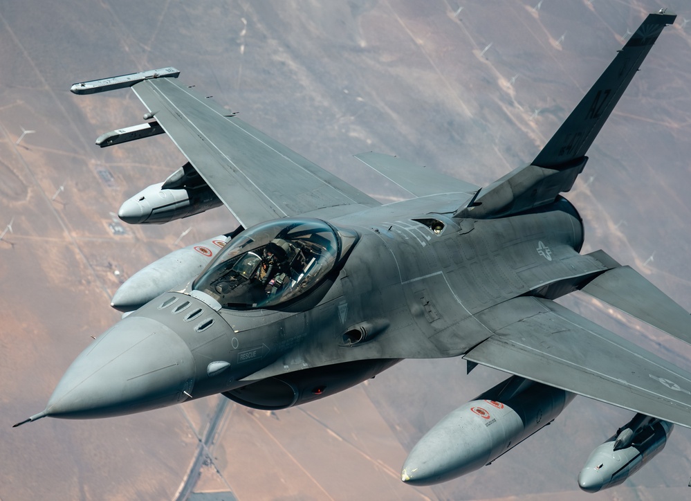 A U.S. Air Force F-16 Fighting Falcon jet fighter is photographed in flight from an aerial perspective, likely from a tanker aircraft during or after aerial refueling. The aircraft is painted in standard gray camouflage and carries multiple external stores on its wing pylons. The terrain visible below appears to be arid desert landscape.